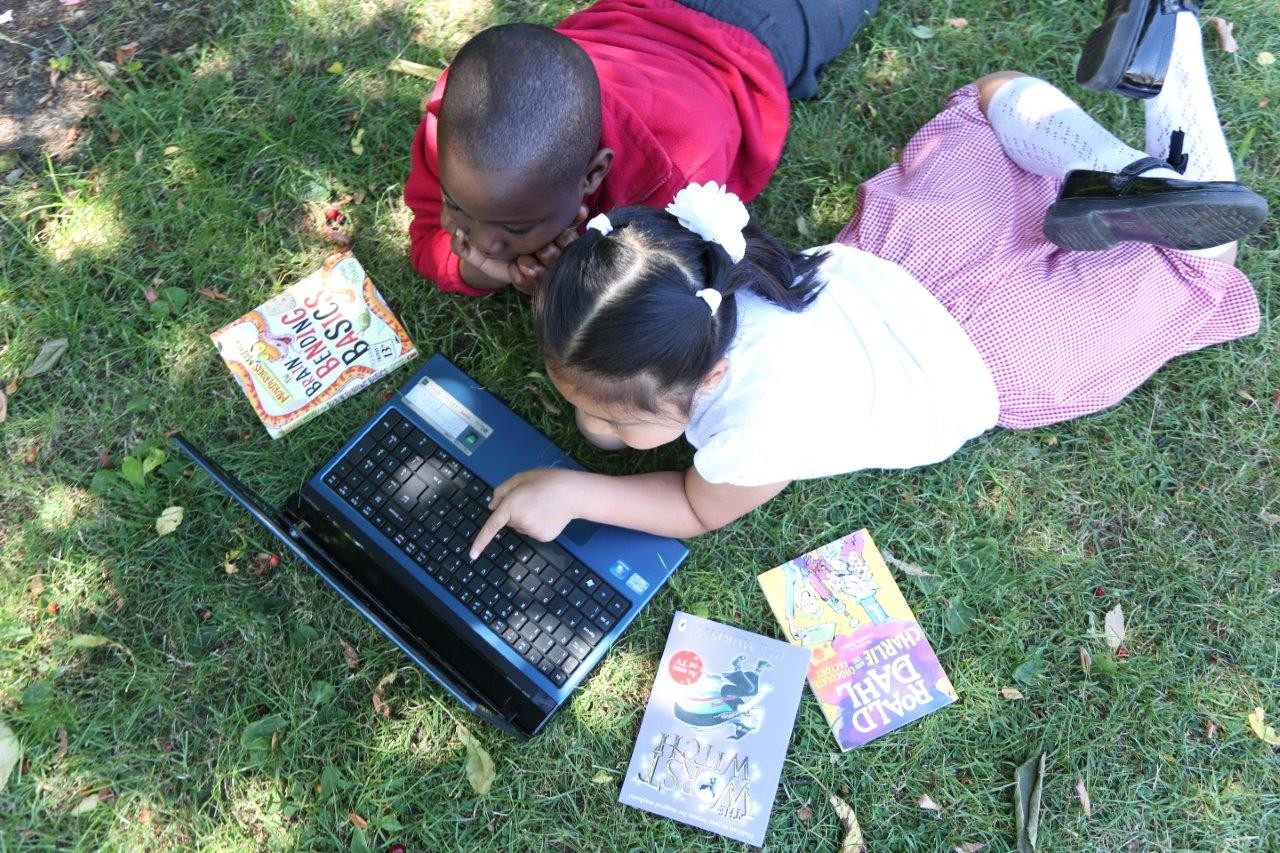 Two children lying on grass looking at a laptop surrounded by paperback books.