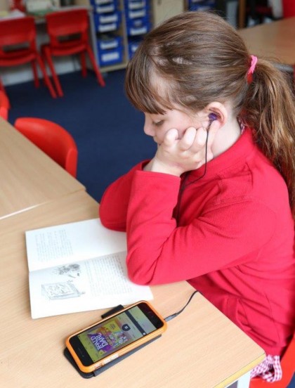 Young girl sitting in a classroom looking a paper book while also listening to it on an MP3 player.