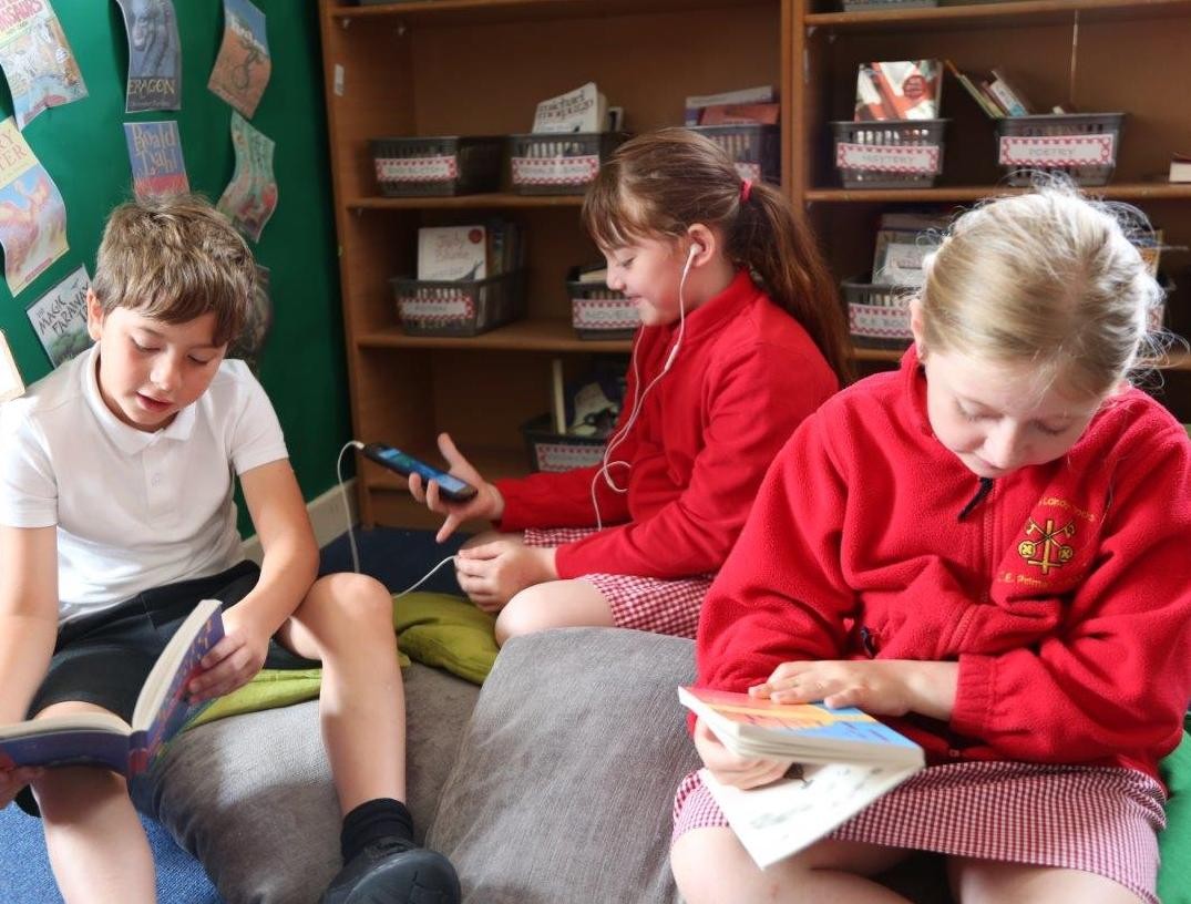 Three children sitting on the floor in a school library. Two children are reading printed books and one is wearing headphones and looking at an iPad.