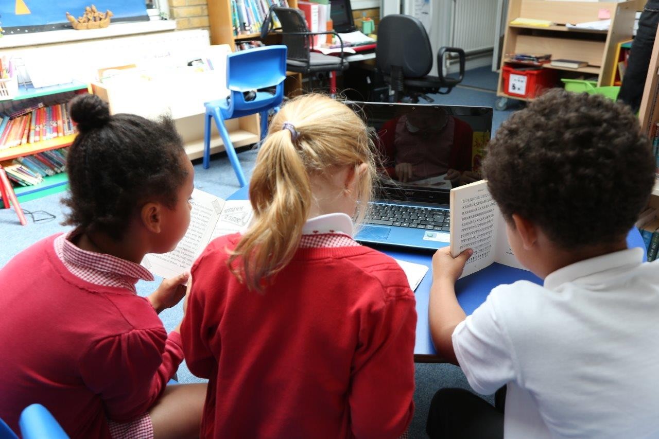 Three children looking at a laptop. One child is reading a printed book.