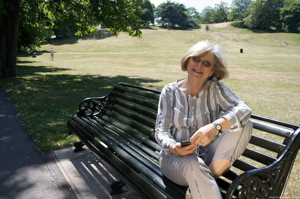 Woman sat on a bench in a park listening to an audiobook on an iPod.