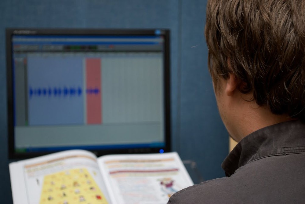 Man sat looking at a computer screen with a sound wave on it, comparing the audio to the book text in front of him.