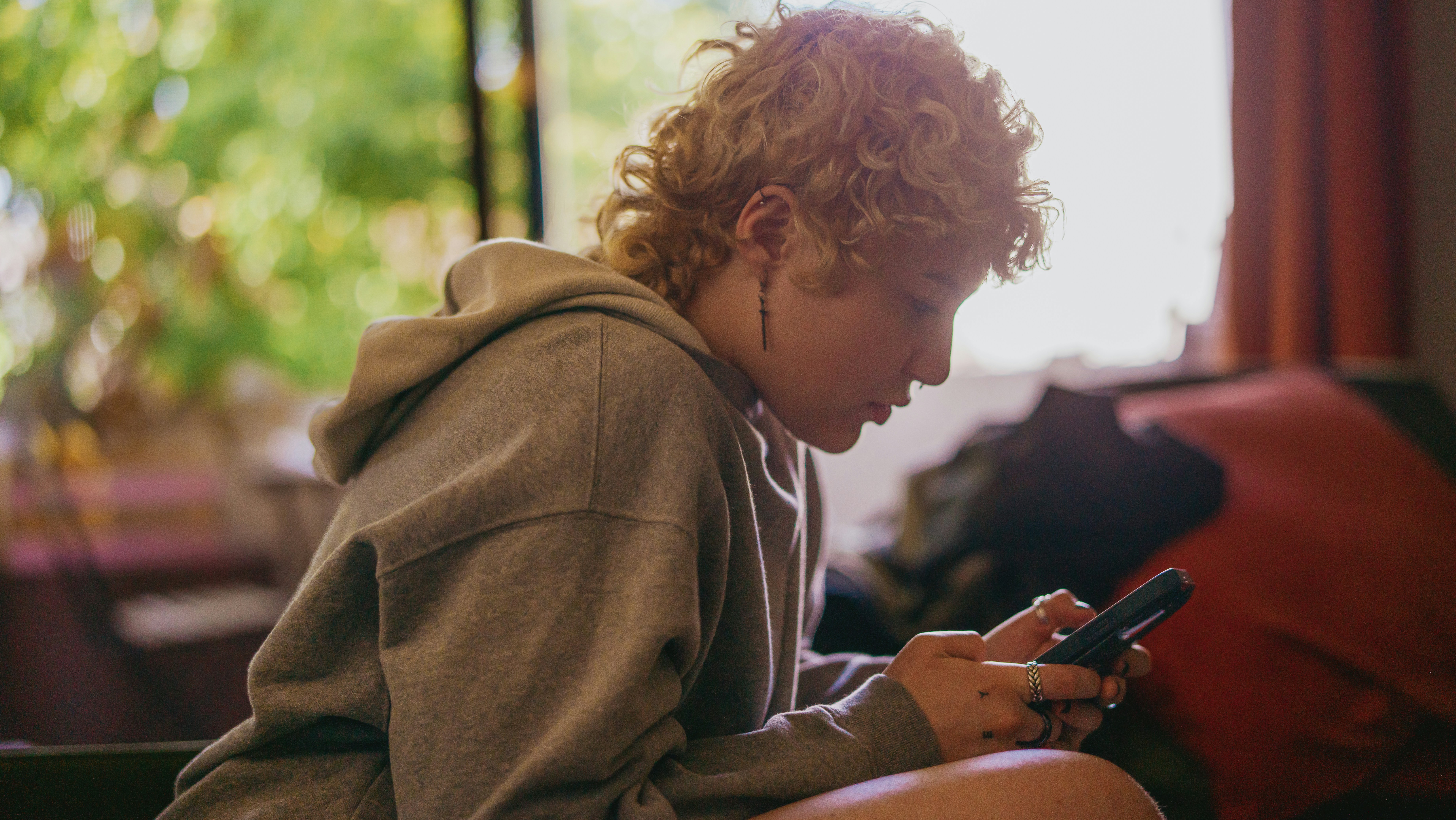 A young person with a grey hoodie is sat looking and engaging with their phone
