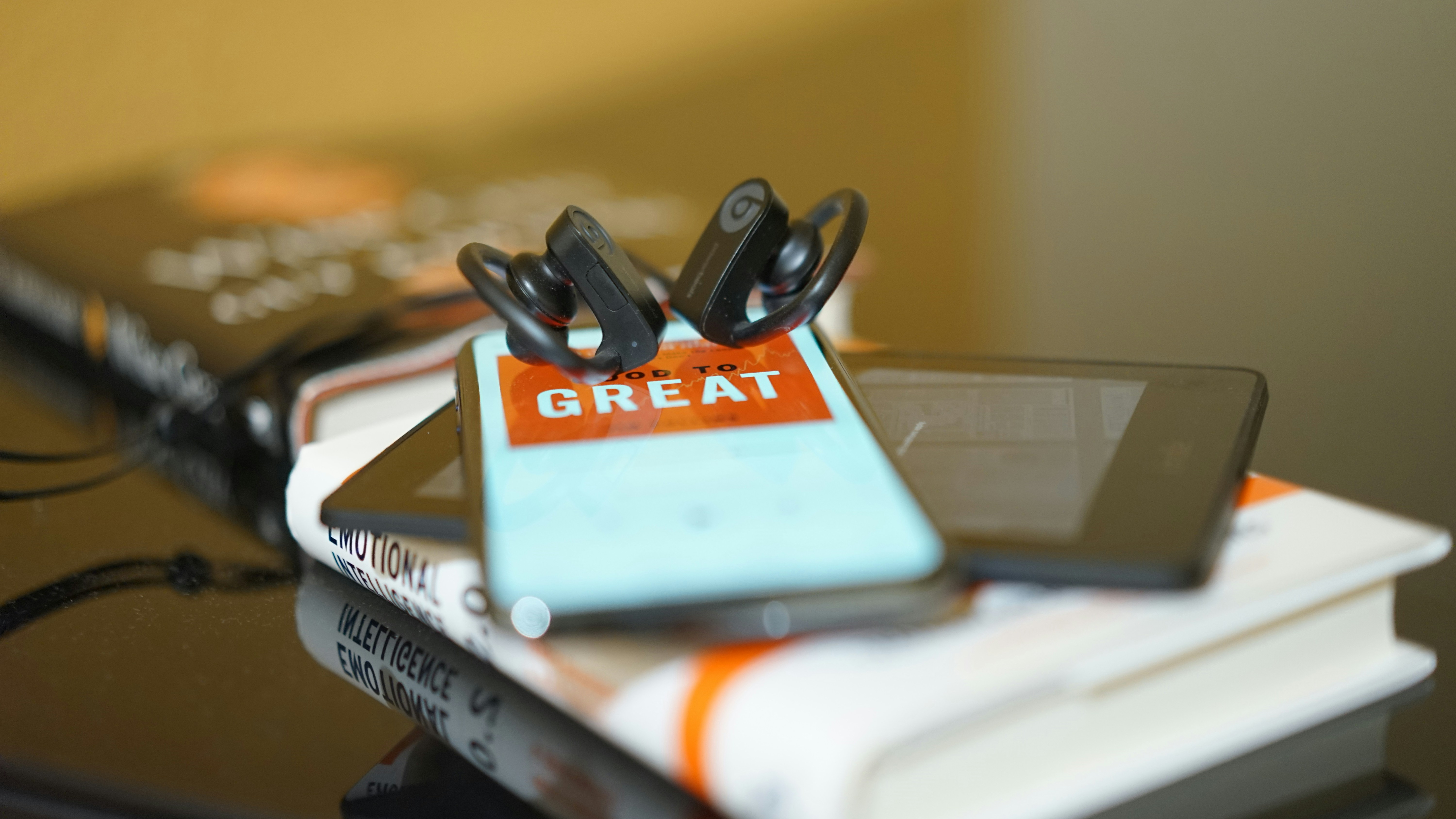 Two black ear phones placed on a phone with audiobook artwork, on top of a kindle and book pile